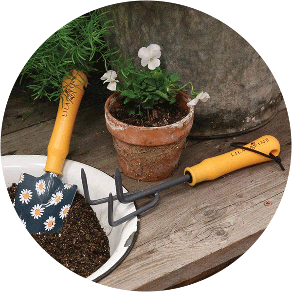 A trowel and gardening fork sitting on a wood table next to some plants and a pot of dirt.